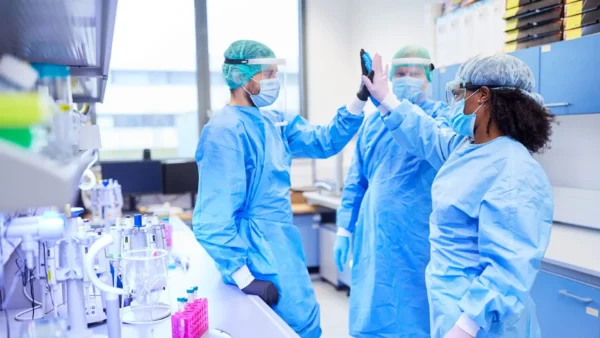 Tres científicos con el EPI completo, incluidas batas, mascarillas y protectores faciales, se chocan los cinco en un laboratorio.